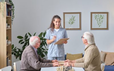 Young nurse making notes in medical card while talking to senior men during their chess play at the table in nursing home