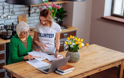 Help elderly people. Beautiful young blond lady volunteering in filling out paperwork for charming elegant silver-haired pensioner in stylish glasses and knitted bright