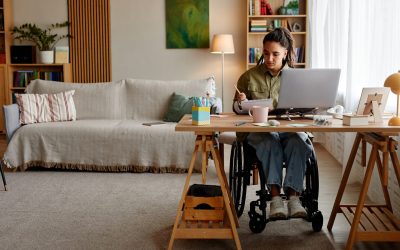 Young adult Caucasian woman with disability sitting in wheelchair working at desk with laptop and notebook in modern living room, focused on writing and studying at home